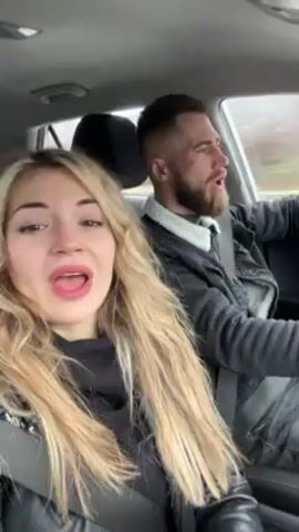 The video shows a couple singing together in a car. The woman is in the passenger seat, wearing a black sweater under a black leather jacket, and has long blonde hair. She is looking at the camera and singing enthusiastically, with a wide open mouth. The man is driving, wearing a black jacket with a light-colored collar, and has a beard and short hair. He is also singing along, looking focused on the road but still participating in the song. The car is moving, and the background through the windshield shows a blurred landscape, possibly on a highway, on a grey day.