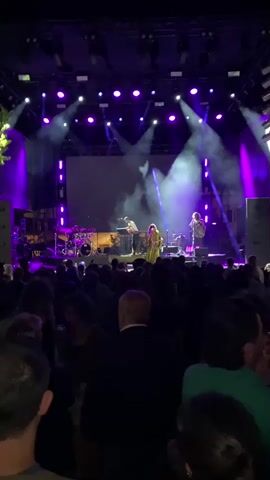 A concert performance is taking place on a dimly lit outdoor stage. A band is performing on stage, with a drummer on the left, a keyboard player in the middle, and two singers in the center and right. The stage is illuminated by purple and white spotlights, and there is a large screen behind the performers. A crowd of people is gathered in front of the stage, watching the performance. The video is shot from the perspective of someone in the audience.
