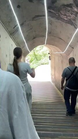 People are walking down a set of wooden stairs inside a concrete tunnel. There are lights on the ceiling of the tunnel. At the end of the tunnel, the people exit into an outdoor area with trees and a bridge.