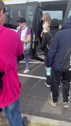 A group of people are boarding a bus. In the foreground, a person wearing a bright pink hooded sweatshirt and a black backpack is facing away from the camera. To their right, another person wearing a dark hoodie and backpack is standing near the bus entrance. A young person with blonde hair, wearing a black outfit, is visible near the bus door, possibly assisting passengers. To the left of the person in the pink hoodie, a person wearing a blue shirt and a camouflage backpack is partially visible. The bus has a yellow stripe along its side and text that appears to read "Komunikacja Gminy Buk". The ground is paved, and the scene appears to be outdoors, possibly at a bus stop or station. The overall action is people boarding a bus.
