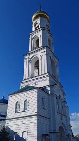 The video shows a close-up, low-angle shot of a white bell tower with a gold dome against a clear blue sky. The tower has several levels with arched openings, some containing bells. A clock face is visible near the top of the tower. The lower portion of the tower is attached to a building with a green roof and arched windows. To the left, a portion of another building with a blue dome is visible. The video is static, with no movement. The lighting suggests it is daytime.