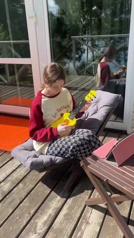 A young woman sits on a lounge chair on a wooden deck playing a yellow ukulele. She wears a white sweatshirt with red sleeves and black and white patterned pants. A laptop with a pink cover sits on a small table to her right. Behind her, large glass doors reflect the outside scenery of trees and sky.