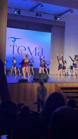 A stage performance is underway, with multiple young dancers dressed in navy blue outfits. One dancer, distinguished in a red outfit, stands out prominently. They are performing in front of a screen displaying the word "Tema" and a logo, indicating this might be the name of the dance club. The stage is well-lit with stage lights, and a set of speakers is positioned in the front. The audience, partially visible in the foreground, watches the performance. One girl stands close to the stage, possibly an audience member or a participant waiting for her turn. The background of the stage includes drawn curtains on either side.