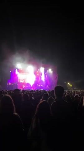 A live music performance takes place at night, filmed from within the audience. The band performs on a large outdoor stage with a screen behind them displaying an image of the lead singer wearing sunglasses and a hooded jacket. The lead singer, sporting a similar look with a colorful hat, stands center stage singing into a microphone. To his left, a guitarist in a baseball jacket plays along. The stage is bathed in bright purple and white light, with smoke effects adding to the atmosphere. The audience, mostly silhouetted against the light, is dense, with some people raising their hands in enjoyment.