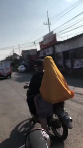 A slightly blurry, medium shot captures two individuals riding a motorcycle on a paved road. The person in the back, presumably female due to the yellow hijab, is wearing a grey skirt and a dark top. The person in front, wearing a dark t-shirt and a dark baseball cap, is driving. The motorcycle has a license plate that is partially visible. The environment appears to be a town or city, with buildings and power lines visible in the background. The sun is out, casting shadows.
