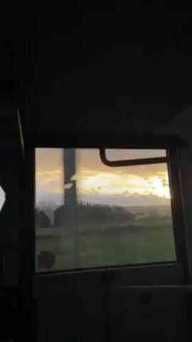 A short video shows a view from inside a vehicle, likely a bus or train, looking out the window. The view transitions from a general landscape of green fields and trees to a close-up of a sky filled with clouds and hints of blue. The sun is setting or rising, creating a warm, golden glow across the sky. The vehicle is moving, as evidenced by the changing scenery. Towards the end, the view passes by trees with bare branches. The interior of the vehicle is dimly lit.