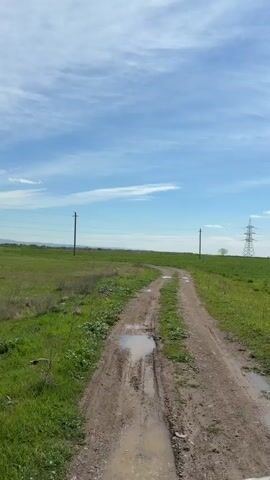 The video shows a dirt road cutting through a grassy field on a sunny day with a few clouds in the blue sky. The road has some puddles, and power lines run along the field with a lattice tower visible in the distance. The camera appears to be low to the ground and stable, and an unidentified sound occurs at the beginning of the video.