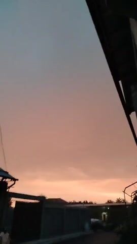 The video shows an outdoor scene at dusk. The sky is a gradient of pink and blue, with clouds visible in the distance. In the foreground, there is a building with a corrugated metal roof and a dark wall. There are also power lines crisscrossing the sky. A person is standing on the left side of the frame, near the building. The ground appears to be paved, and there is a fence or wall behind the building. The overall impression is one of a quiet, residential area at the end of the day.
