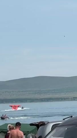 A jet ski tows a red inflatable water toy across a body of water. The toy is shaped like an upside-down T with several seats or sections. The jet ski is creating a wake behind it, and the inflatable toy is lifting out of the water. In the foreground, the tops of several people's heads and shoulders are visible, along with parts of parked cars and a green tarp. In the background, rolling green hills are visible under a clear blue sky.
