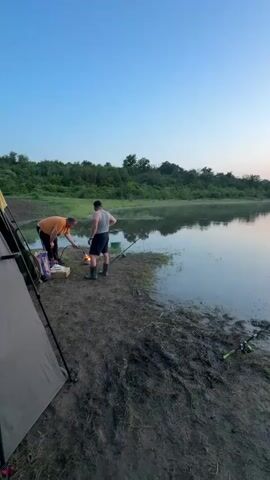 Two men are camping and fishing by a tranquil lake at dawn. One man, in an orange shirt, is kneeling beside a camping stove and a box, appearing to set something up. The other man, in a white tank top and shorts, stands beside him, observing. Two fishing rods are set up along the muddy bank, their lines cast into the calm water. A tent is partially visible on the left side of the frame. The background features lush green trees and a clear sky, with the rising sun casting soft colors across the horizon, reflected beautifully in the water. The scene suggests a peaceful morning fishing trip.