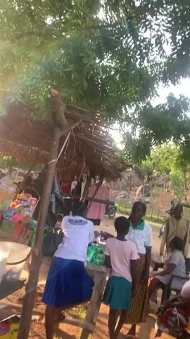 The video shows an outdoor scene with several people gathered near a makeshift structure with a thatched roof. The structure appears to be a stall or a small shop, with items displayed on tables and hanging from the roof. In the foreground, a person wearing a white t-shirt with "CONSOLIDATION 2020" printed on it is interacting with another person wearing a pink t-shirt. Behind them, several other individuals are visible, some standing and some seated. A blue motorcycle is parked to the right of the frame. The background consists of trees, rocks, and a dirt ground. The overall setting suggests a rural or semi-rural environment. The lighting indicates it is daytime, possibly late afternoon.