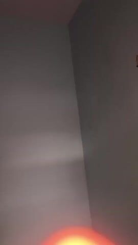 The video displays a dimly lit room. The camera pans across walls, showing a light fixture and what appears to be a light switch on one wall. There are a few dark smudges or marks on the walls, some of which appear to be in focus and have a reddish-brown hue, while others are out of focus. The lighting suggests it may be nighttime or the room is not well-lit.