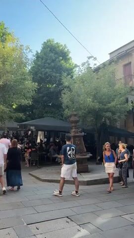 The video shows a street scene with several people walking and sitting at outdoor tables under umbrellas. The paving stones are gray and patterned. There are trees and buildings lining the street. A fountain is visible on the right side of the frame. The people are dressed in casual summer clothing. The sky is blue and clear, indicating a sunny day. Some people are walking towards the camera, while others are walking away. Some are in groups, and some are alone. There is a general sense of pedestrian activity and outdoor dining.