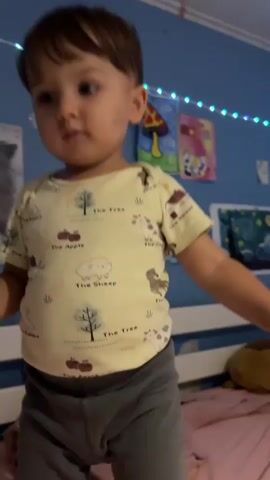 A young child with brown hair and dark eyes is standing in front of a blue wall decorated with drawings and fairy lights. The child is wearing a light yellow onesie with a pattern of trees, rabbits, and apples, and grey pants. The child is holding a small box of purple objects in their left hand and looking down. A person's hand reaches into the frame from the right and appears to be interacting with the child or an object the child is holding. The child then turns their head to the left, and the person's finger touches the child's chin. The child looks surprised or curious. The background includes a white bed frame and a pink bedspread. The video appears to be shot indoors.