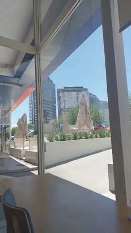 The video shows an outdoor scene viewed through a window. Outside the window, there's a paved area with a white wall. Behind the wall, there are several closed, beige-colored patio umbrellas, along with some greenery. In the background, there are tall buildings under a clear blue sky. A red car is parked in the distance. Inside, near the window, there is a table and a chair. The table is the most prominent object in the foreground. The camera zooms in slightly on the exterior view.