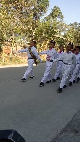 A group of young people in white uniforms with red sashes are marching or performing a synchronized routine outdoors on a paved surface. They are wearing dark caps and white gloves. The person in the foreground is holding a large, flat object with a golden edge, possibly a prop or a flag. There are trees and some buildings in the background. The sun appears to be shining.