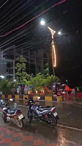 The video shows a street scene at night with rain falling. In the foreground, two motorcycles are parked on the side of the road. A streetlight with an unusual, elongated illuminated fixture hangs overhead. Beyond the motorcycles, there are trees and buildings. A few people and a rickshaw are visible in the background, moving through the rain. The road surface is wet and reflective.