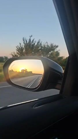 The video shows a side-view mirror reflecting a road and a sunset. The sun is low on the horizon, casting a warm glow over the landscape. Trees and fields are visible on either side of the road, and the sky is a clear blue. The camera appears to be inside a car, looking out the window at the mirror.