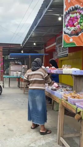 The video shows an outdoor market stall. In the foreground, a person wearing a hijab and a striped sweater over a denim skirt is browsing various food items displayed on wooden tables. Another person in a hijab is visible behind the counter, potentially serving customers. The stall has a bright orange banner with colorful food illustrations and text in Indonesian. The market appears to be situated along a street, with a parked motorcycle and a yellow truck visible in the background. The sky is overcast.