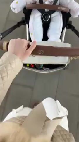 A first-person perspective shows a person pushing a baby stroller with a white diaper bag attached to the handlebar. The baby is strapped in, wearing a white snowsuit with pink and yellow star patterns. The diaper bag has a light brown handle and is partially open, revealing items inside. The person pushing the stroller is wearing a beige coat and a quilted white bag hanging from their shoulder. The stroller moves across a tiled sidewalk. The stroller is a Cybex brand.