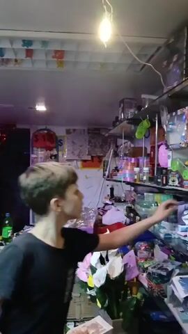 A young boy with short brown hair is standing in front of shelves packed with various items. He wears a black t-shirt and reaches toward the shelves with his right hand. The shelves are made of glass and are filled with colorful ribbons, toys, containers, and other miscellaneous objects. The environment appears cluttered and possibly a shop or storage area. In the background, there are posters, signs with price tags, and additional storage. The lighting is somewhat dim and fluorescent. The overall impression is one of a busy, disorganized space.