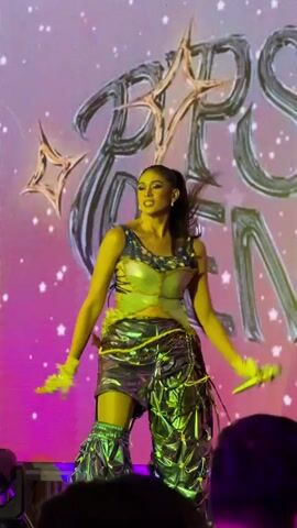 The video shows a person in a unique outfit on a stage with a large screen behind them. The person is wearing a metallic silver outfit consisting of a halter top and baggy pants with intricate detailing and embellishments. They also have on matching metallic gloves. The person is holding a small, light-colored object in their hands. The screen behind them displays a large, stylized graphic with star-like elements and text that is partially obscured. The stage is lit with purple and yellow lights, and some stage equipment is visible at the bottom of the frame. The person is facing towards the audience and appears to be performing or posing.