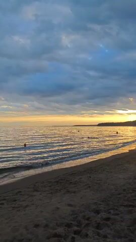 The video shows a beach scene at dusk. The sky is filled with dramatic, cloudy formations with streaks of orange and yellow light from the setting sun visible on the horizon. Gentle waves lap onto a sandy shore, creating a peaceful atmosphere. Several orange buoys are visible in the water, marking a boundary or a designated area. The overall environment is calm and serene, with no people or animals present in the frame. The lighting suggests the end of the day, with shadows beginning to lengthen across the beach.