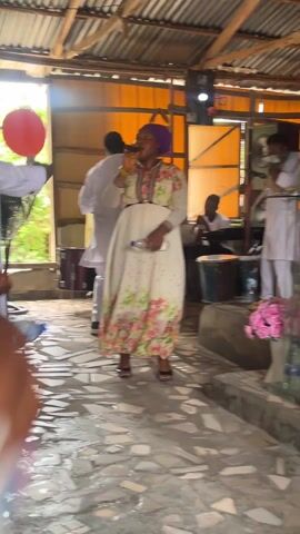 The camera circles around the interior of a small church, revealing a group of people engaged in what appears to be a celebratory service. There is a singer, backed by a keyboard player and drummer, and dancers wearing white, one holding a red balloon. Other churchgoers stand watching or joining in the dancing. A large, stationary electric fan is visible. The space has a corrugated metal roof with exposed wooden beams. The walls are light orange, and one wall behind the stage is covered in purple fabric. The floor is paved with irregular stone tiles.
