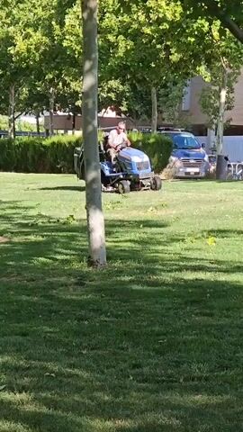 A person is operating a blue lawnmower in a grassy area. The lawnmower is a rider mower with a small trailer attached to the back. The person is wearing a light-colored shirt and pants, and sunglasses. The person is looking forward as they operate the mower. The lawn is green and appears to be well-maintained. In the background, there are trees, bushes, and a building. There is also a blue van parked in the background. The sun is shining, casting shadows on the grass. The scene appears to be in a park or a residential area.