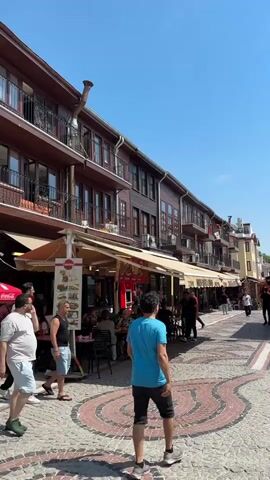 A daytime, eye-level, slightly upward-tilted shot shows a crowded street with people walking and dining at outdoor restaurants. The street is paved with light-colored cobblestones and features a decorative red and brown patterned path in the center. On the left side, multi-story buildings with dark brown wooden facades and numerous windows line the street. Many of these buildings have awnings extending over outdoor seating areas where patrons are visible. A prominent, curved, metal pipe extends from the side of one building. On the right, the buildings are less uniform in color and style. Further down the street, on the right, there are more outdoor dining setups with red canopies. A man in a blue shirt and black shorts walks away from the camera in the center of the street. To his left, several men walk towards the camera, including one in a light gray t-shirt and light blue shorts who is talking on a phone, and another in a black t-shirt and dark pants. The sky above is clear and bright blue.