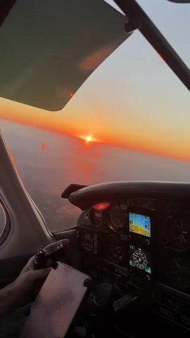 The video shows the interior of a small airplane's cockpit during sunset. The sun is visible on the horizon, casting an orange glow across the sky and reflecting on the water below. The pilot's hand is visible on the yoke. The cockpit instruments are illuminated, including a GPS display. The plane appears to be in flight.