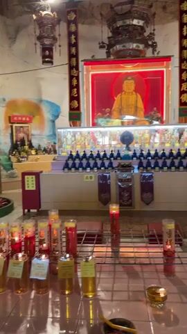 This video is a walkthrough inside a temple. The scene opens with a close-up on a shelf containing several Buddha statues. The camera then pans to the right, revealing a large statue of a female figure adorned in a shimmering robe, standing on a lotus flower. Further to the right, a row of smaller Buddha statues sits illuminated on red platforms, with a mural of clouds and a sunset visible behind them. The temple's interior features stone walls and a mosaic tiled floor. Red lanterns are hung from the ceiling. Several candles and offerings are placed on a metal grate in the foreground.