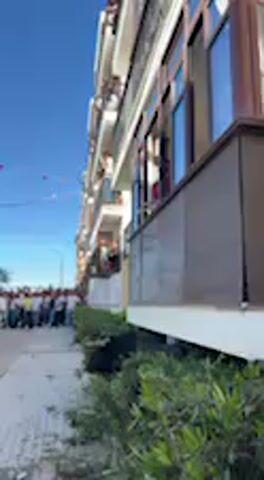This video captures a vibrant daytime scene in Vejer de la Frontera, Spain, featuring a lively street lined with multi-story residential buildings. The architecture is characterized by balconies with brown awnings or sun blinds and numerous windows, reflecting the bright blue sky. Green bushes line the foreground of the street, while colorful bunting or flags are strung across the road in the distance, suggesting a festive occasion.

A large crowd of people is visible on the street, gathered as if for an event or celebration. Several individuals are also observing the scene from the balconies of the buildings. In the immediate foreground, a person's hand is visible, gesturing towards the buildings, seemingly presenting or pointing out features of the scene. The weather is clear and sunny, indicating a pleasant day. No discernible text is visible in the frame. The overall atmosphere appears communal and celebratory.