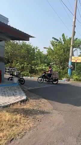 A person on a motorcycle is driving on a paved road. The motorcycle has a cage attached to the back and the person is wearing a red shirt and a mask. There are trees and foliage in the background. A bicycle is parked to the left of the road. The sky is blue and clear.
