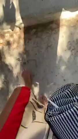 The video shows a first-person perspective of a person walking on a sandy surface. The person is wearing white pants with a red trim and a colorful pattern. The person is barefoot. There are shadows from trees or other objects on the ground. A blue and white striped bag is also visible. The ground appears to be mostly sand with some sparse vegetation.