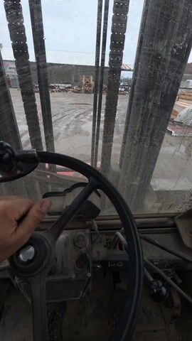 The video shows a first-person perspective from inside what appears to be a heavy machinery vehicle, like a forklift. A person's hand is visible on the steering wheel, with the thumb briefly raised in a thumbs-up gesture. The vehicle's dashboard is visible, with various controls and gauges. Through the dirty windshield and the vertical bars in front, a construction or industrial yard is seen, featuring stacks of lumber, muddy ground, and other pieces of equipment or structures. The sky is overcast.