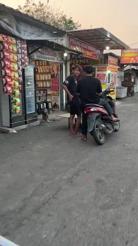 Two young males are standing and interacting outdoors in front of a row of shops. The male on the left is wearing a black t-shirt and black shorts and is facing the other male. The male on the right is sitting on a red scooter, facing away from the camera, and wearing a black t-shirt and dark shorts. The scooter is parked on a paved surface. Behind the males are several stalls or shops with goods displayed, including packaged items and what appear to be bottled liquids. One sign reads "PISANG KEJU SUKODONO". The environment appears to be an outdoor market or street scene with a slightly overcast sky.