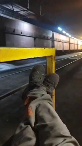 The video shows a pair of work boots and legs, resting on a yellow railing. The person is wearing grey work pants with orange accents and dark work boots. In the background, a long train with many cars sits on railroad tracks. The scene is dimly lit, with artificial lights illuminating the train cars and the surrounding industrial area. The overall impression is one of a break or pause during a shift in an industrial setting.