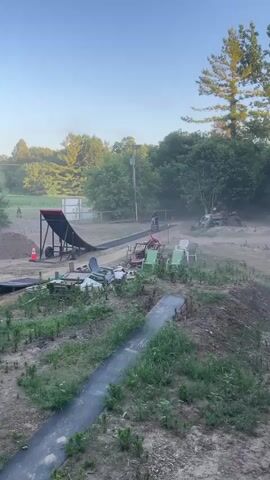 The video shows a motocross rider performing a flip over a ramp. The rider is wearing a black helmet and black gear. The motorcycle is blue and black. The rider launches off a dirt ramp and flips in the air. The rider then lands on an inflatable airbag. The video is shot from a slightly elevated angle. The environment appears to be an outdoor motocross track with trees and a blue sky in the background. There are also some chairs and other debris scattered around the track.