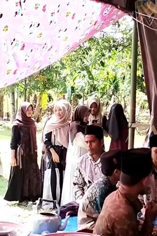 The video shows a celebration of the Prophet Muhammad’s birthday, known as Maulid Nabi Muhammad. There are several people present, including men in traditional Muslim caps and attire, women wearing hijabs, and several children. Some of the children are performing a clapping exercise, likely as part of the celebration. A sign in the background announces the event. The celebration appears to be taking place in an outdoor or semi-outdoor setting, with a canopy providing shade and shelter.