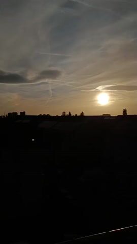 The video displays a sunset over a cityscape. The sun is a bright orb in the upper right portion of the frame, casting a warm glow on the sky. Wispy clouds stretch across the sky, some appearing as thin streaks and others as thicker, darker formations. The horizon is lined with the silhouettes of buildings and trees, all rendered dark against the illuminated sky. The foreground is almost entirely black, obscuring any details of the immediate surroundings, but a faint railing or ledge is visible at the very bottom of the frame. The overall impression is one of twilight descending upon an urban landscape.