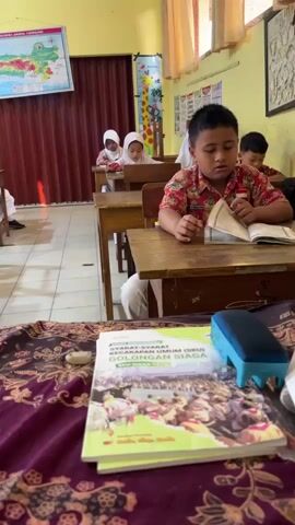 The video shows a classroom scene with several young children seated at wooden desks. The children appear to be students, and they are mostly facing forward, engaged in an activity. One boy in the foreground is looking down at an open book on his desk, holding a pen. He is wearing a red and white patterned shirt. Several girls wearing white hijabs are seated further back in the classroom, also looking at their desks and books. The classroom walls are yellow, and there are maps and charts displayed on the walls. A dark red accordion-style door is visible on the left side of the room. In the foreground, a book with a green cover is placed on a table covered with a dark red patterned cloth. The overall atmosphere suggests an educational setting, possibly a lesson or study period.