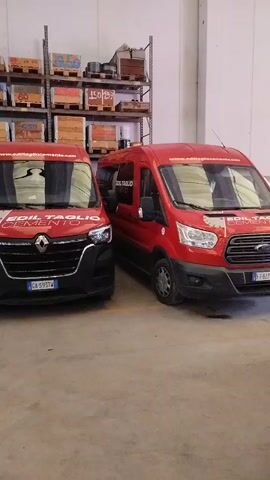 This video captures a scene inside a warehouse or garage in Villapinta, Italy. The primary subjects are several red vans, parked in rows. Two vans are prominently featured at the beginning, one facing forward and another angled. Further into the video, more red vans are visible, including one exiting the warehouse through a large open garage door. The warehouse has concrete floors and walls, with shelves stacked with boxes visible in the background. The garage door is open, revealing an outdoor scene with greenery and mountains, suggesting a daytime setting. The lighting appears to be artificial, typical of an indoor industrial space. No people are visible in the footage.

Visible text on the vans includes "COIL TAGLIO" and what appears to be a logo with horizontal lines. Another van has text that is partially obscured but includes what looks like "CIOPLANO ELEMENTO". The overall impression is one of an organized vehicle fleet within a commercial or industrial facility.
