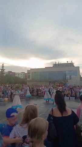 This is a vertical video clip filmed at an outdoor event in Krivodanovka, Russia. The scene is set in the late afternoon or early evening, with a cloudy sky indicating potential for rain or just overcast conditions. A group of young girls in traditional-looking dresses with white tops and light blue skirts are performing a dance in the center of a paved area. They are wearing wreaths of greenery on their heads.

A large crowd of spectators is gathered around the dancing area, watching the performance. The people in the foreground, mostly out of focus, appear to be families with children, suggesting a community event or festival. The backdrop includes buildings, some of which appear to be residential blocks, and a larger, modern-looking building with a glass facade. There is also a lone pine tree visible to the left.

The overall atmosphere seems festive and engaging, with the focus on the cultural performance. The people watching appear to be enjoying the spectacle. No discernible text is visible in the video.