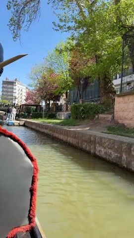 The video captures a scenic gondola ride on a canal in a city, likely Eskisehir, Turkey, as suggested by the context.

**Subjects:** The main subjects are a male gondolier, dressed in a blue jacket, jeans, and a cap, steering the gondola with a long pole, and two female passengers. One passenger wears a black cap and glasses, while the other wears sunglasses. Other people are visible walking along the canal bank in the background.

**Objects:** The prominent objects include the black gondola with red trim, the gondolier's long oar, and the canal itself. Along the canal banks, there are multi-story brick and lighter-colored buildings, some appearing to be apartments and others storefronts. Lush green trees, some with pink or red blossoms, line the banks. A bridge can be seen in the distance, and the water in the canal is a greenish-brown color.

**Activity/Event:** The video shows a leisure activity, a gondola tour along the canal. The gondolier is actively propelling the boat, while the passengers are looking around, taking in the sights. One of the passengers is holding a smartphone.

**Notable Details:** The scene takes place on a bright, sunny day under a clear blue sky, suggesting pleasant weather for an outdoor activity. The green foliage and blossoms indicate spring or early summer. The buildings and general urban setting create a lively city atmosphere.

**Visible Text:** The brand name "Penti" is clearly visible on a building along the canal, indicating a shop.