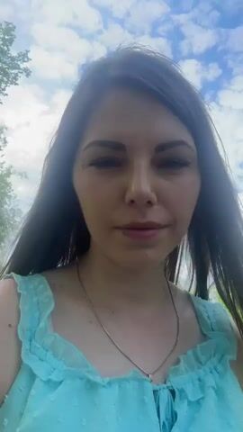 A woman with long dark hair, wearing a light blue dress and a thin gold necklace, is holding the camera and filming herself from a close-up perspective. She is standing on a dirt path bordered by green grass and trees. The sky above is partly cloudy. The camera is fairly stable, and the woman maintains eye contact with the lens. Her expression is neutral.