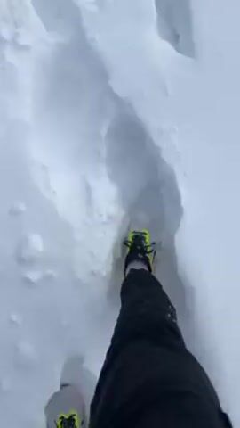 A first-person perspective shows someone walking in deep snow. The person is wearing black pants and light-colored running shoes with bright yellow and black accents. The depth of the snow is apparent, as each step creates a large impression. The snow is pristine and white, and the lighting suggests a bright, possibly overcast day. The person appears to be moving steadily through the snow, navigating the uneven terrain.