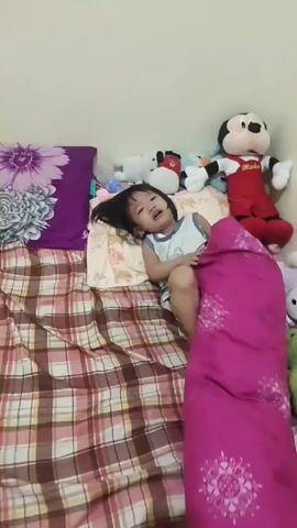 A child is lying on a bed covered by a pink blanket. The child is holding the blanket over their face and appears to be playing. Several stuffed animals are visible on the bed behind the child.