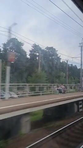 A view from a train window shows a train platform in a wooded area. The platform has a white railing, benches, trash cans, and a signpost. Several people are visible on the platform, some standing and one sitting on a bench. Overhead power lines are visible against a cloudy sky. The train tracks are in the foreground.