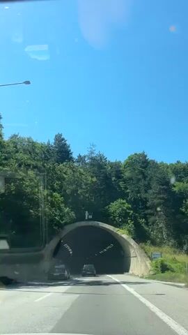 The video captures a point-of-view shot from inside a vehicle traveling through a tunnel. The initial shot shows the bright, sunlit exterior of the tunnel entrance surrounded by green trees. As the vehicle enters the tunnel, the scene transitions to darkness, illuminated by rows of lights along the ceiling and walls. Other vehicles can be seen ahead. The road surface appears to be wet in some areas. The video ends as the vehicle continues deeper into the tunnel.